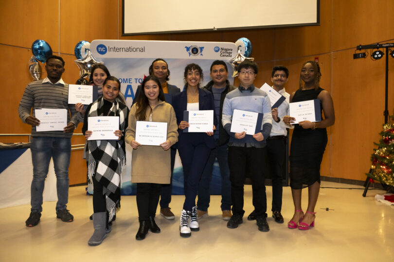 A group of students holding award certificates standing in front of a sign that reads 'NC International'.