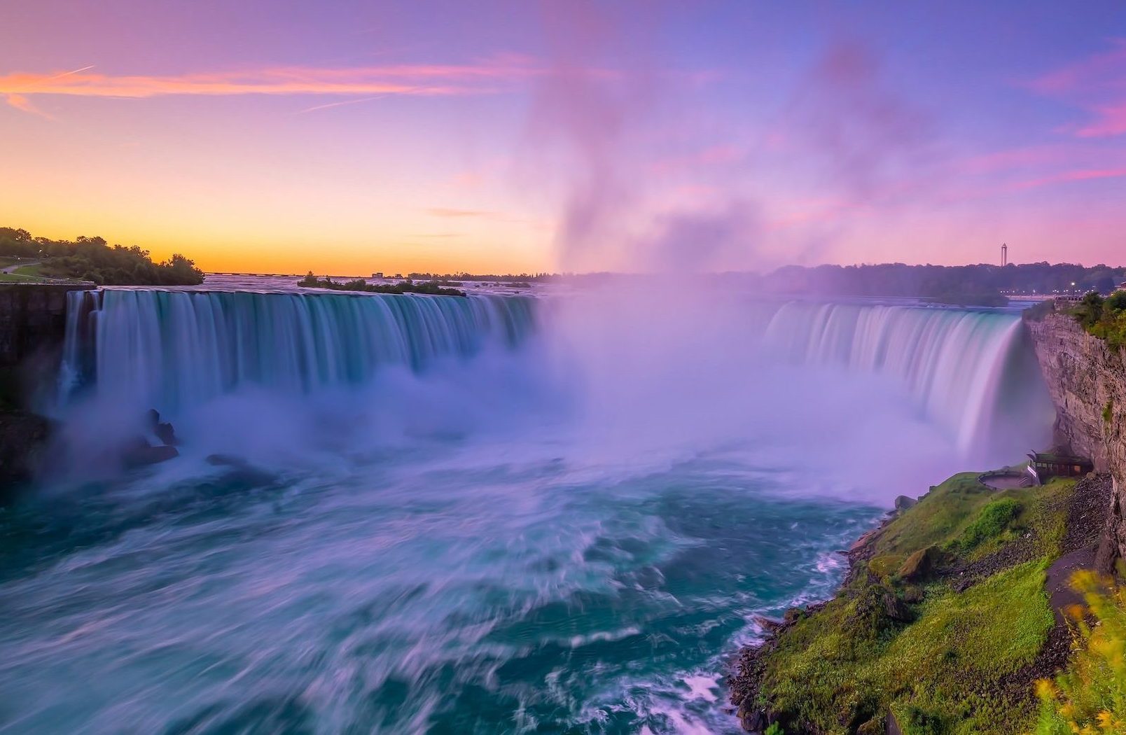 A wide view of Niagara Falls at sunrise, with water flowing over the cliffs and mist drifting up into the sky.