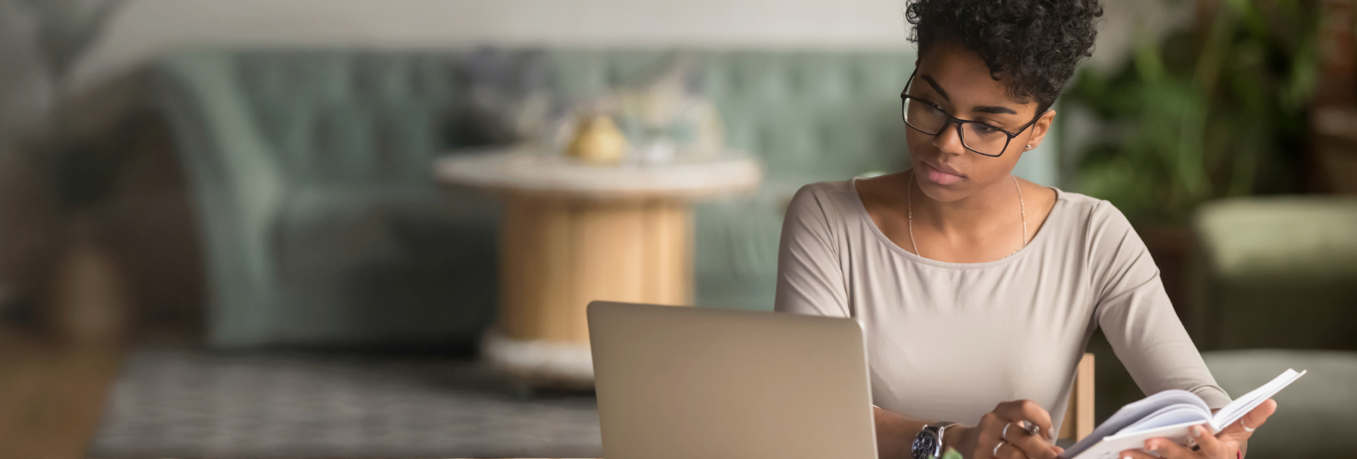 Individual wearing glasses sits at a table, looking at a laptop and writing in a notebook.