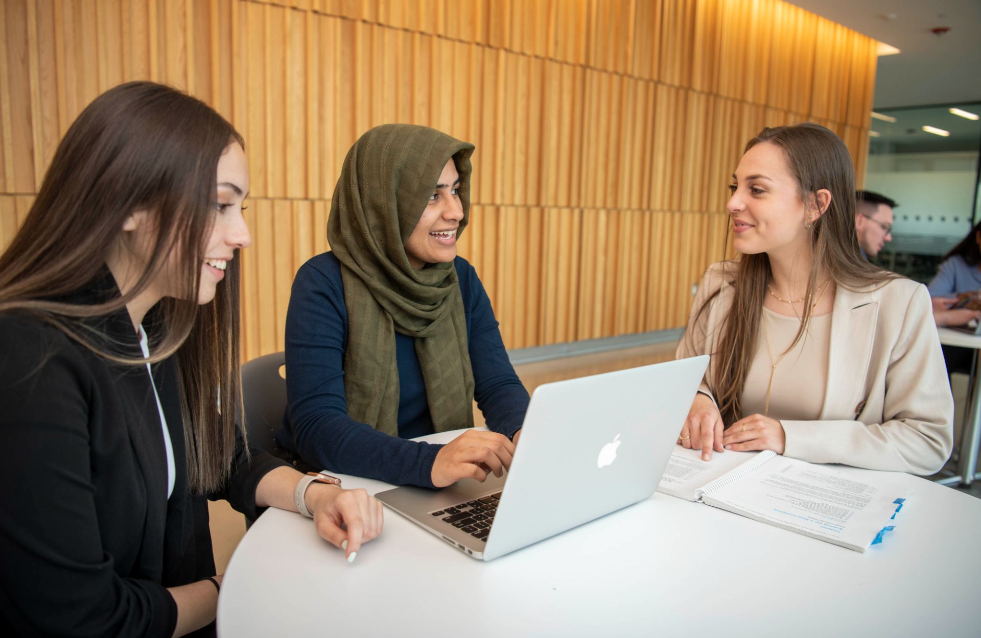 Three people sit at a round table, collaborating with a laptop and documents in a modern, wood-panelled room.