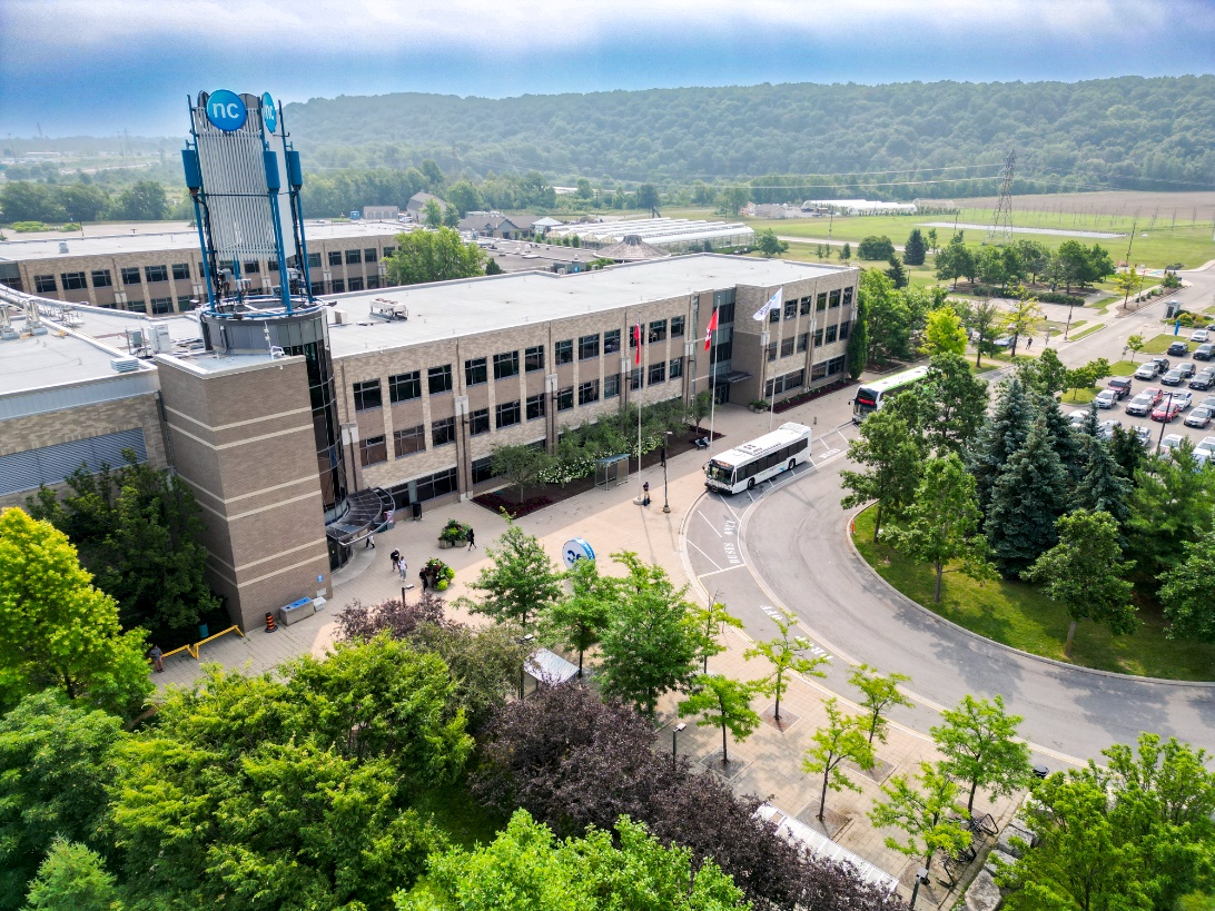 Aerial view of a large campus building with a bus at the entrance, surrounded by green spaces and car parks.