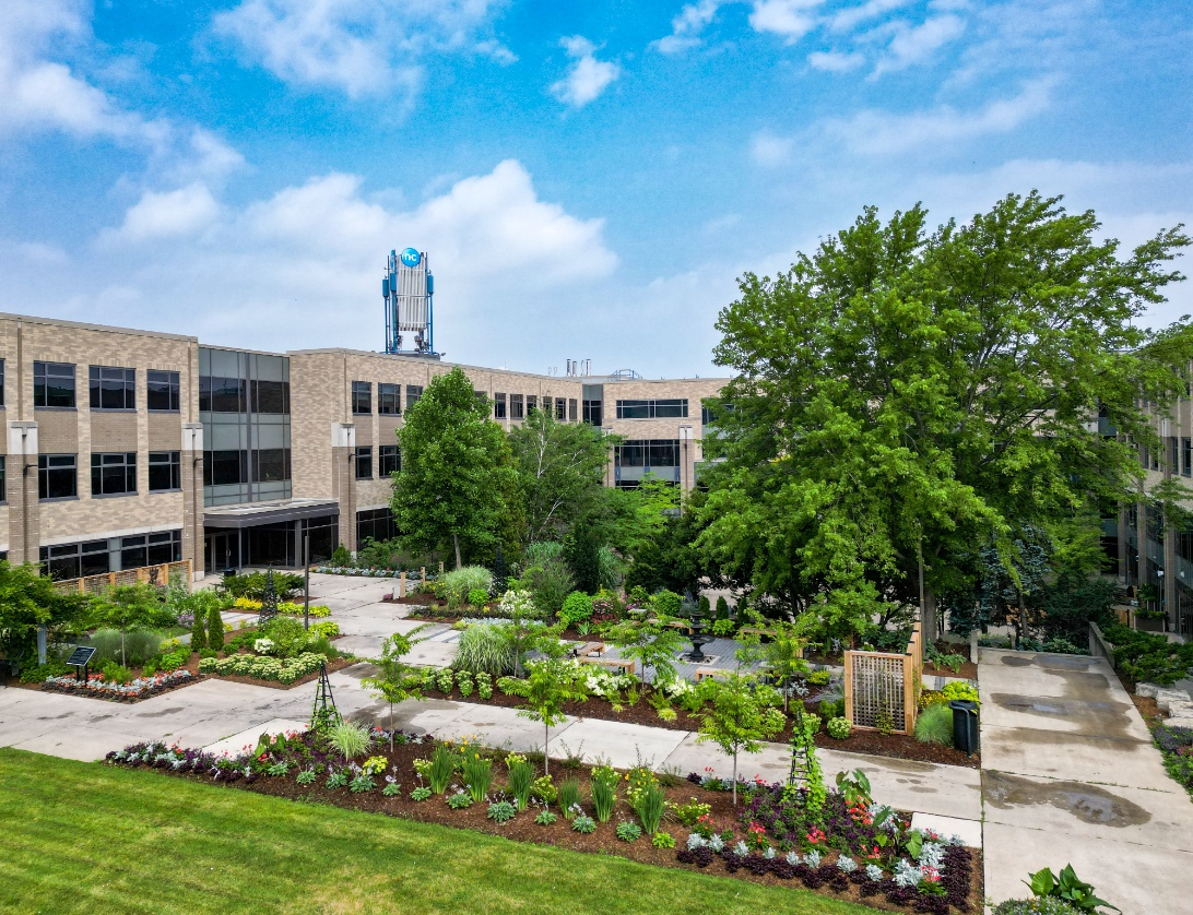 A modern three-storey building featuring a landscaped courtyard, gardens, and trees under a partly cloudy sky.