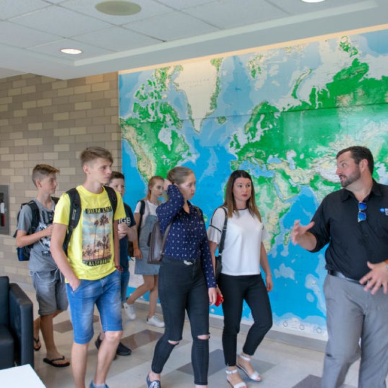 A group of students listens to a person speaking in front of a large world map displayed on a wall.