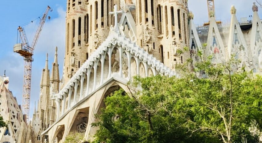 Front view of the Sagrada Familia basilica in Barcelona, with a construction crane and green trees visible in the foreground.