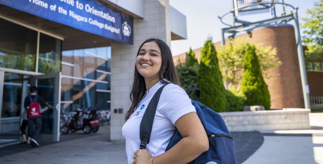 A student wearing a rucksack stands and smiles outside a Niagara College building during orientation.