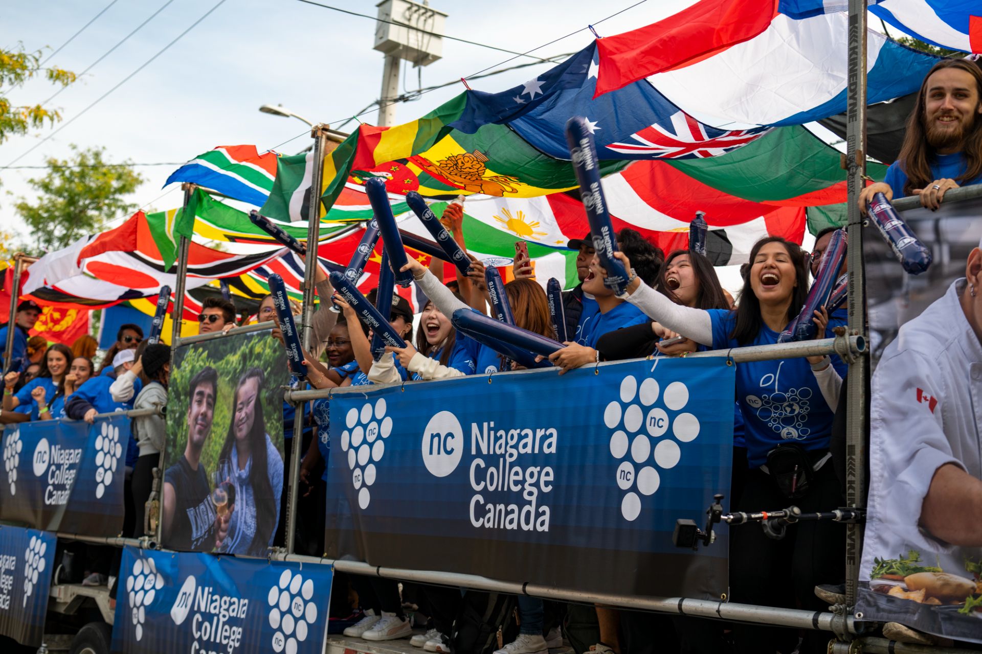 A group holding blue Niagara College Canada banners cheers near a display of various international flags.
