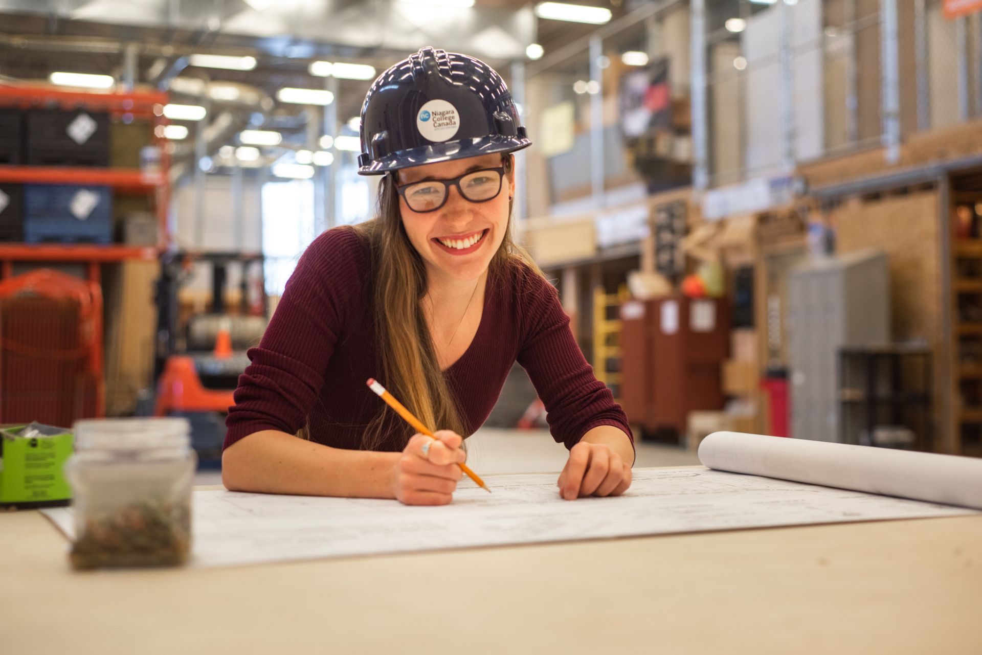 A person wearing a hard hat and glasses smiles whilst working on architectural plans in an industrial workspace.