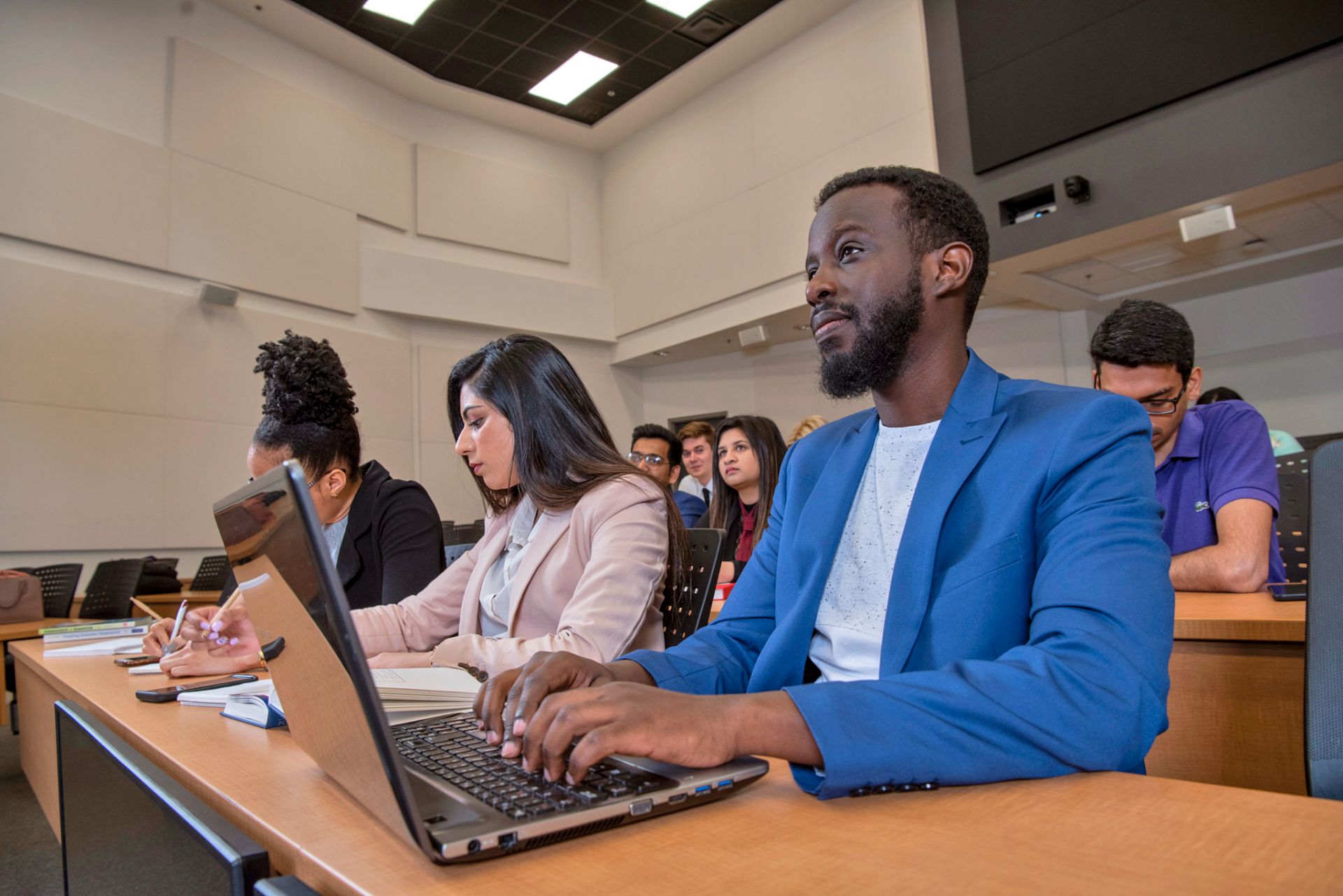 Several students are seated at desks in a classroom, taking notes and typing on laptops during a lecture.