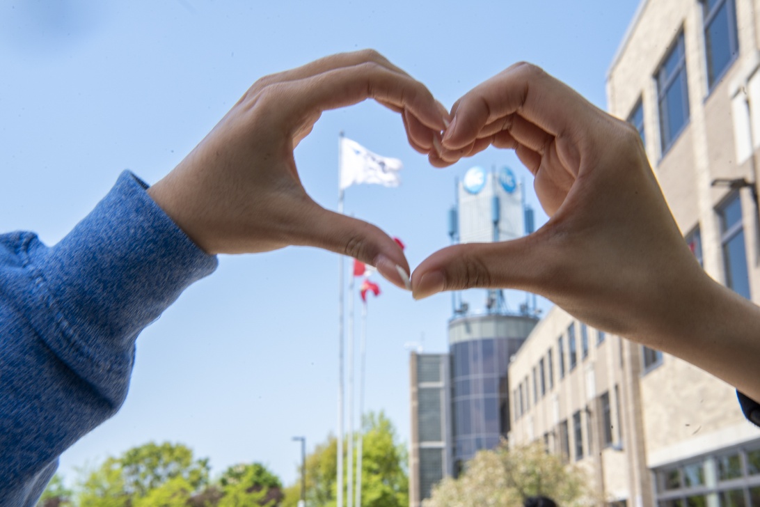 Hands create a heart shape in the foreground, with flags and a modern building seen in the background.