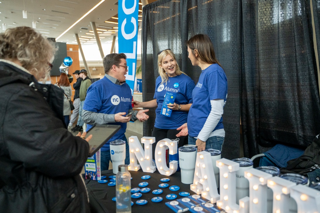 Three individuals in blue "NC Alumni" shirts chat at a stall displaying NC Alumni signs, badges, and cups.