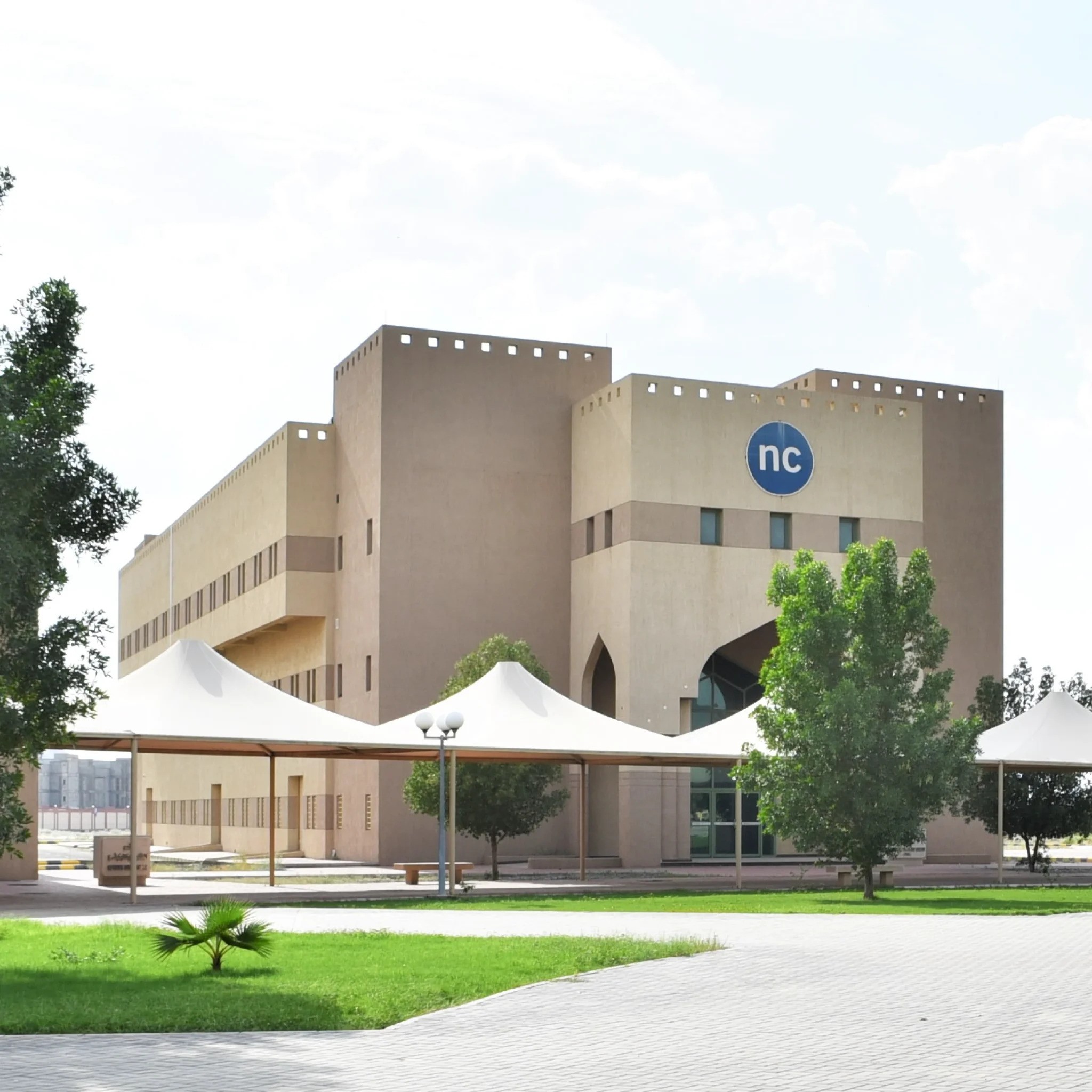 A modern beige building featuring an "nc" logo, set among trees, grass, and white shade structures.
