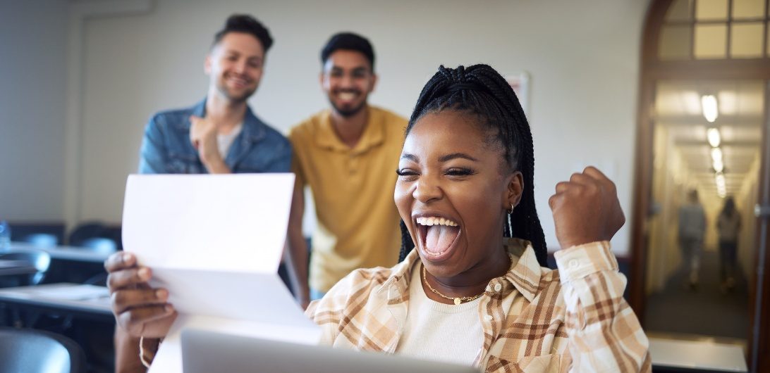 A person sits at a desk, smiling and raising their fist whilst holding a paper, with two people standing behind them.
