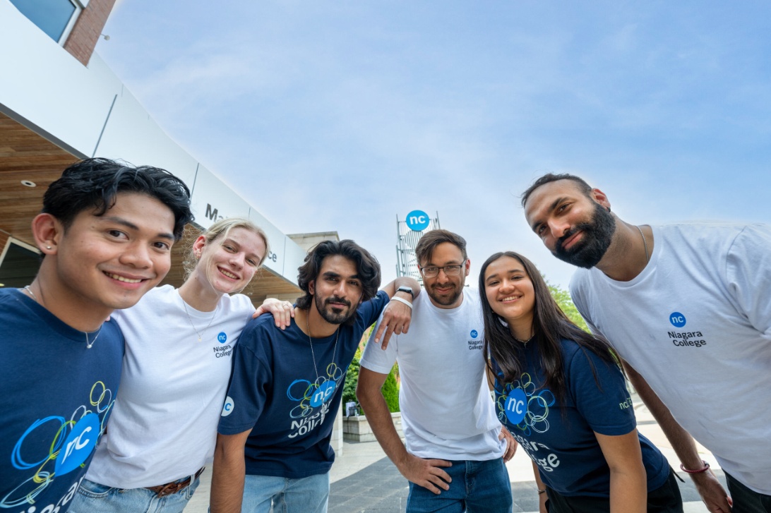 Six Niagara College students pose and smile outside near a campus building on a sunny day.