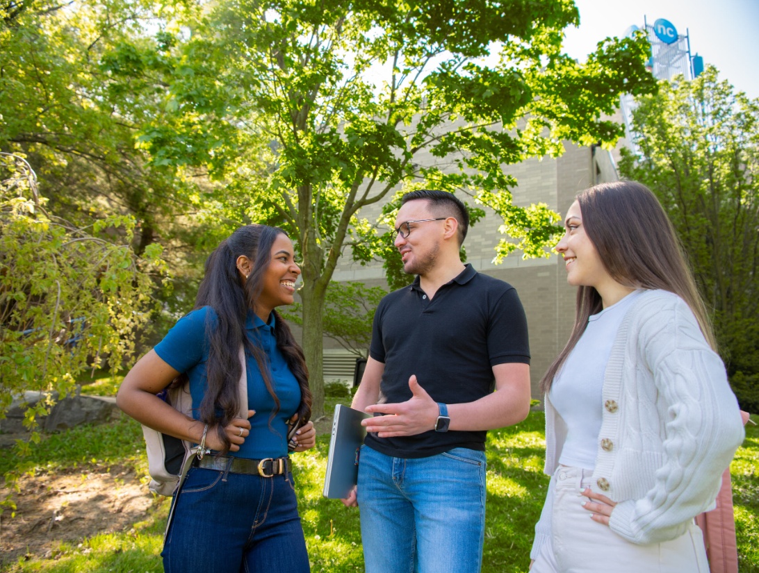 Three people stand outside on grass, talking and smiling, with trees and a building in the background.