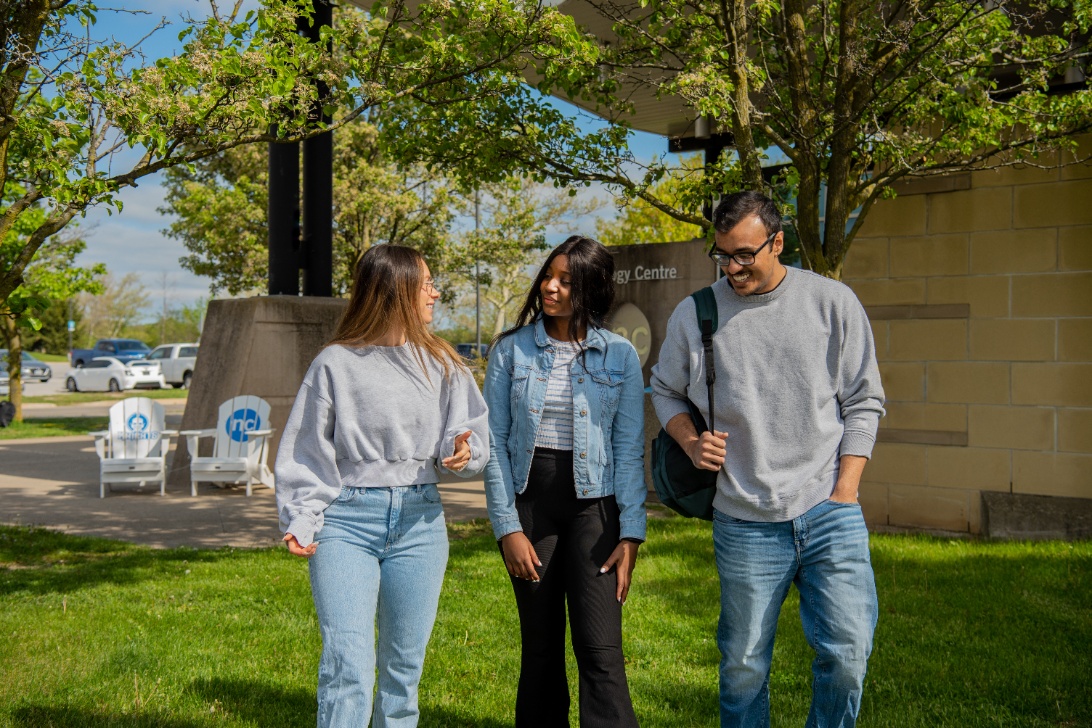 Three people walk together on a grassy area outside, chatting and smiling, with trees and a building in the background.