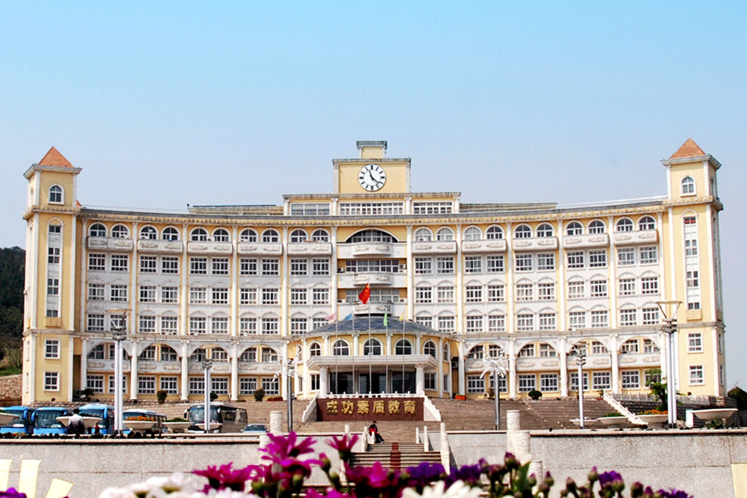 Large, multi-storey building with a clock on top, central entrance, flags, steps, and flowers visible from the front view.