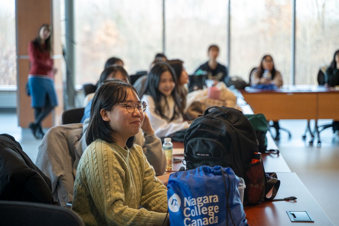 Students sit at desks in a classroom; a student in front has a Niagara College Canada bag on the table.