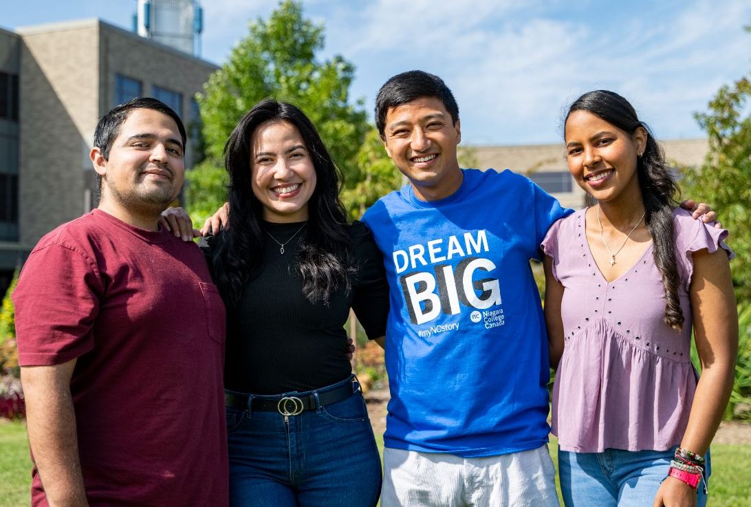 Four young people stand close together outside, smiling at the camera on a sunny day.