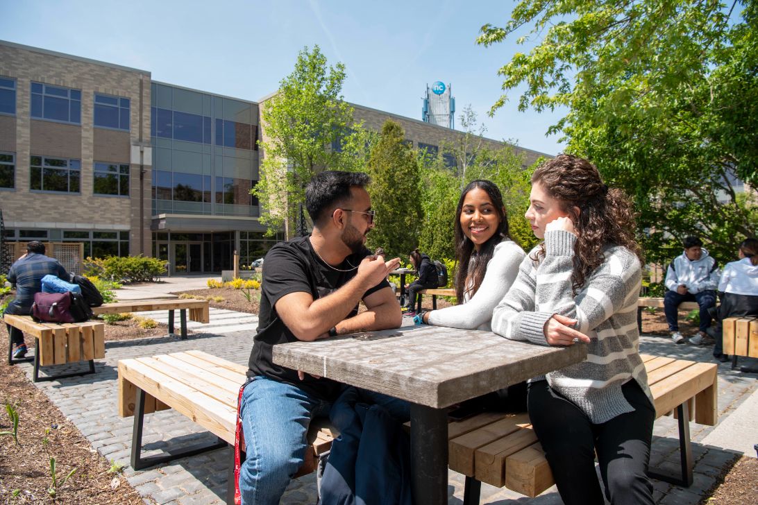 Three people sit and chat at an outdoor table on a sunny day in front of a modern campus building.