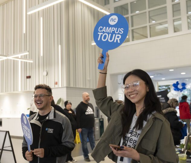 Two individuals indoors hold "Campus Tour" signs, smiling; more people are visible in the background of a modern building.
