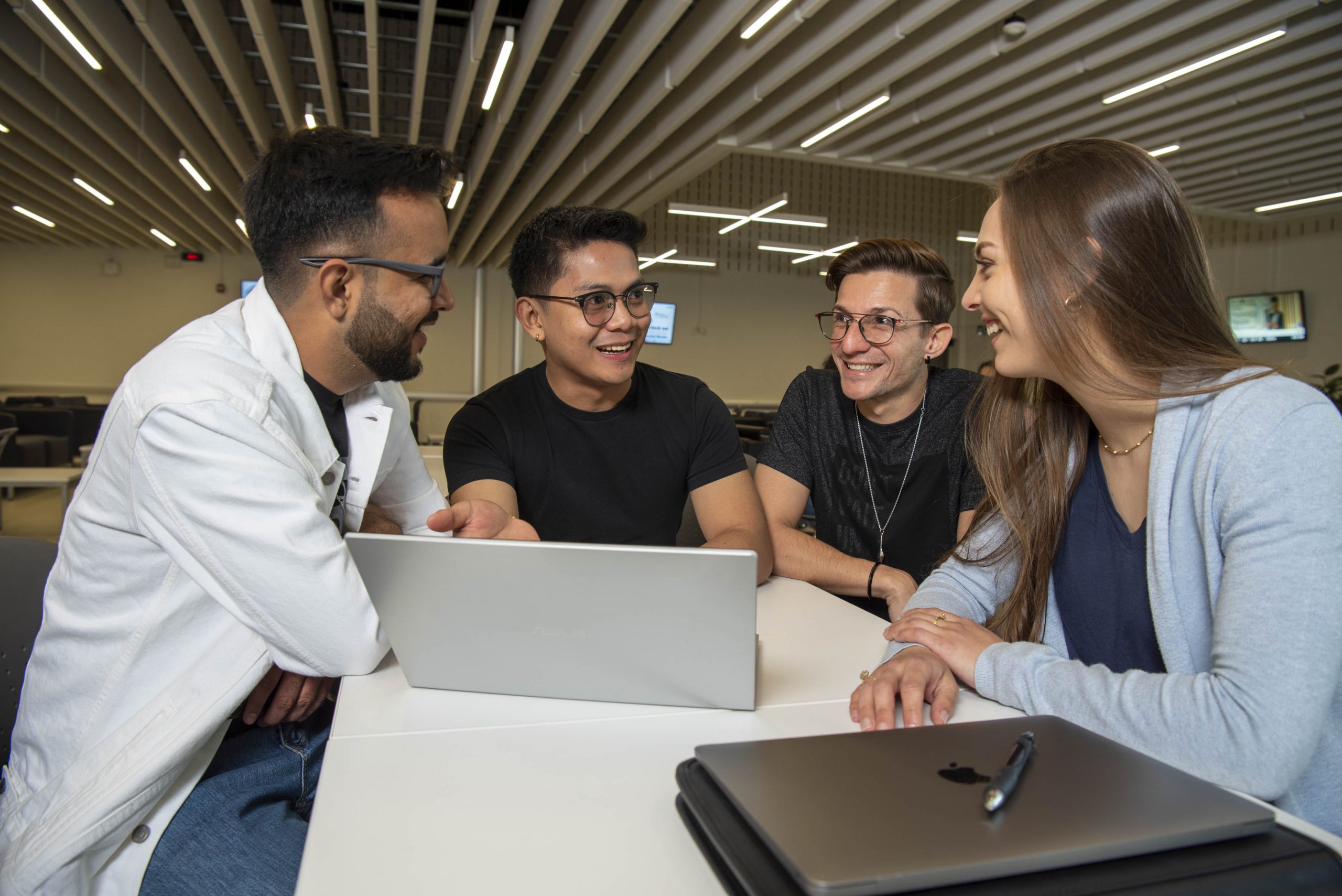 Four people sit at a table with laptops, smiling and chatting in a modern, well-lit room.