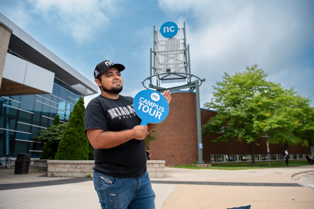 Someone holds an "NC Campus Tour" sign outside a modern college building and tower featuring the NC logo.