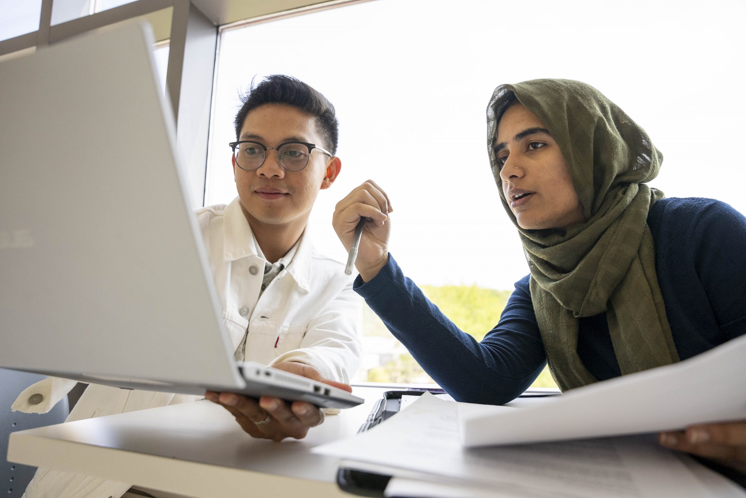 Two individuals sit at a table, viewing a laptop screen as they discuss documents and take notes.