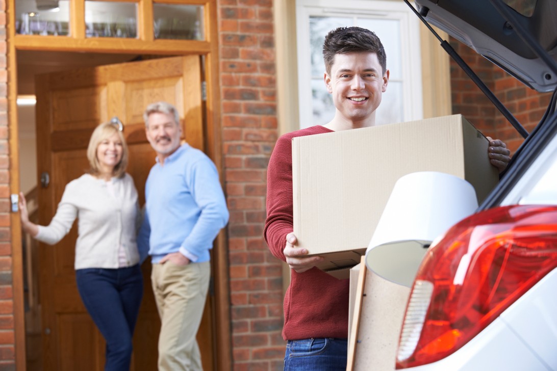 Young person carrying a cardboard box from a car, with two adults standing and smiling by a house entrance in the background.