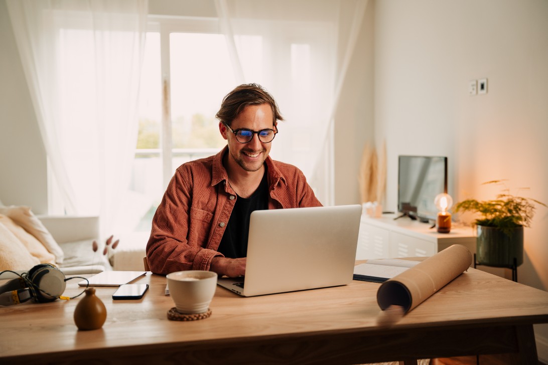 Person wearing glasses works on a laptop at a wooden desk in a bright, modern home office with natural light.