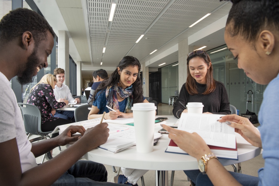Four students sit at a table in a modern building, reading and writing with books and coffee cups placed in front of them.