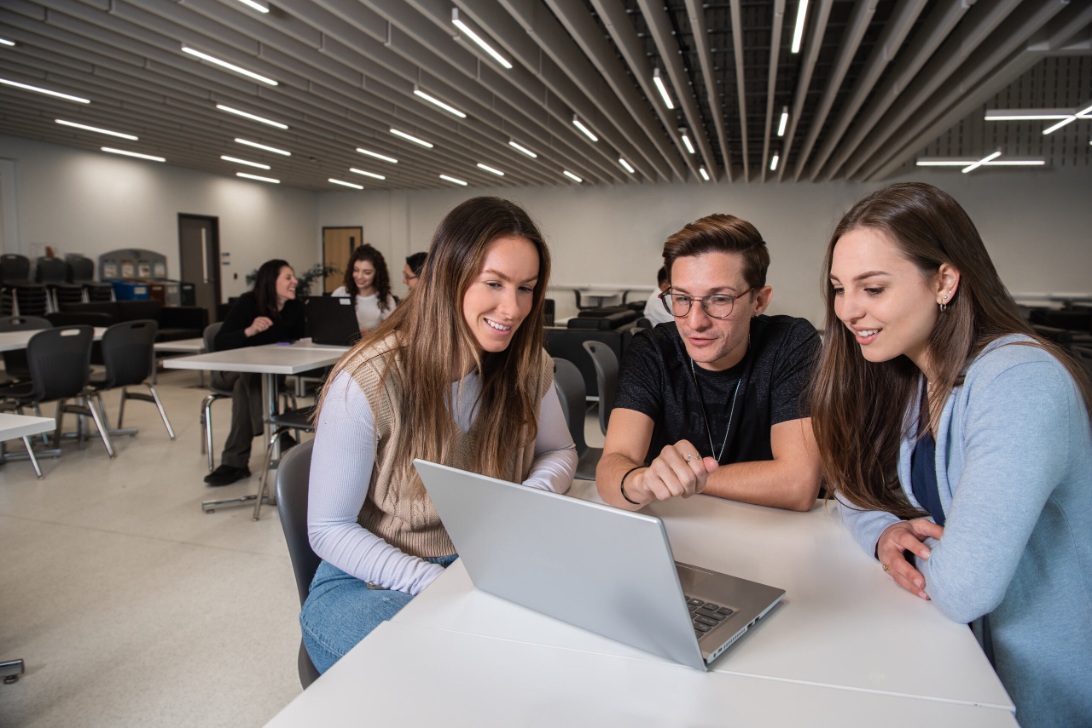 Three young people sit at a table looking at a laptop in a modern classroom with other individuals in the background.