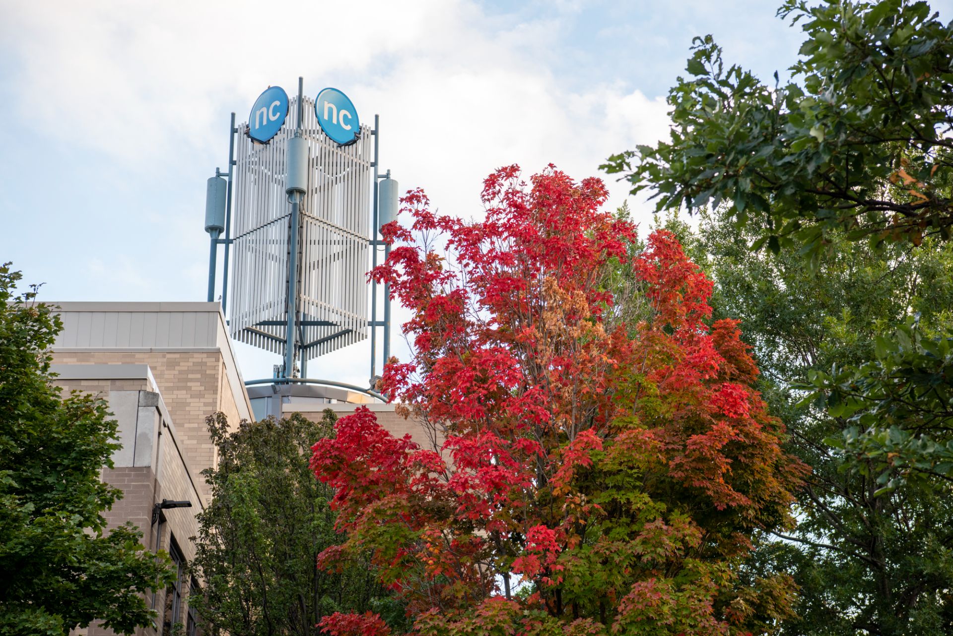 A building displaying "NC" signage with a tall tower rises behind a tree with red autumn leaves under a partly cloudy sky.