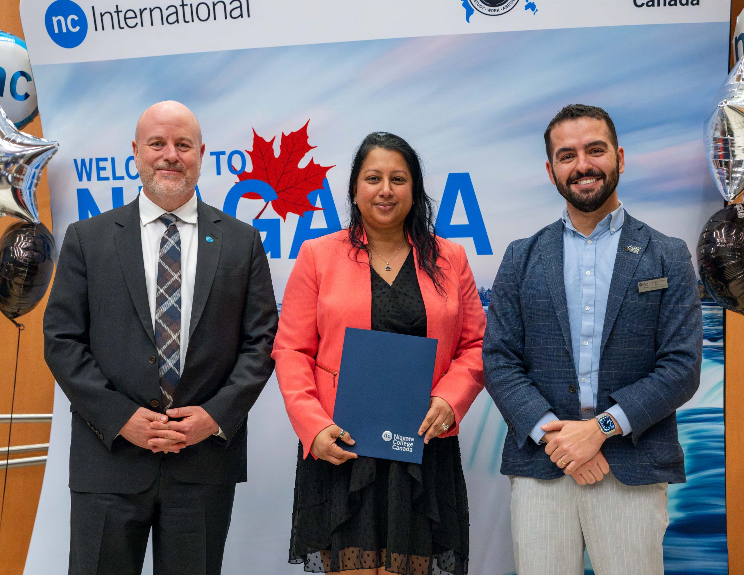 Three individuals standing and posing for a photo at a Niagara College International event, one holding a folder.