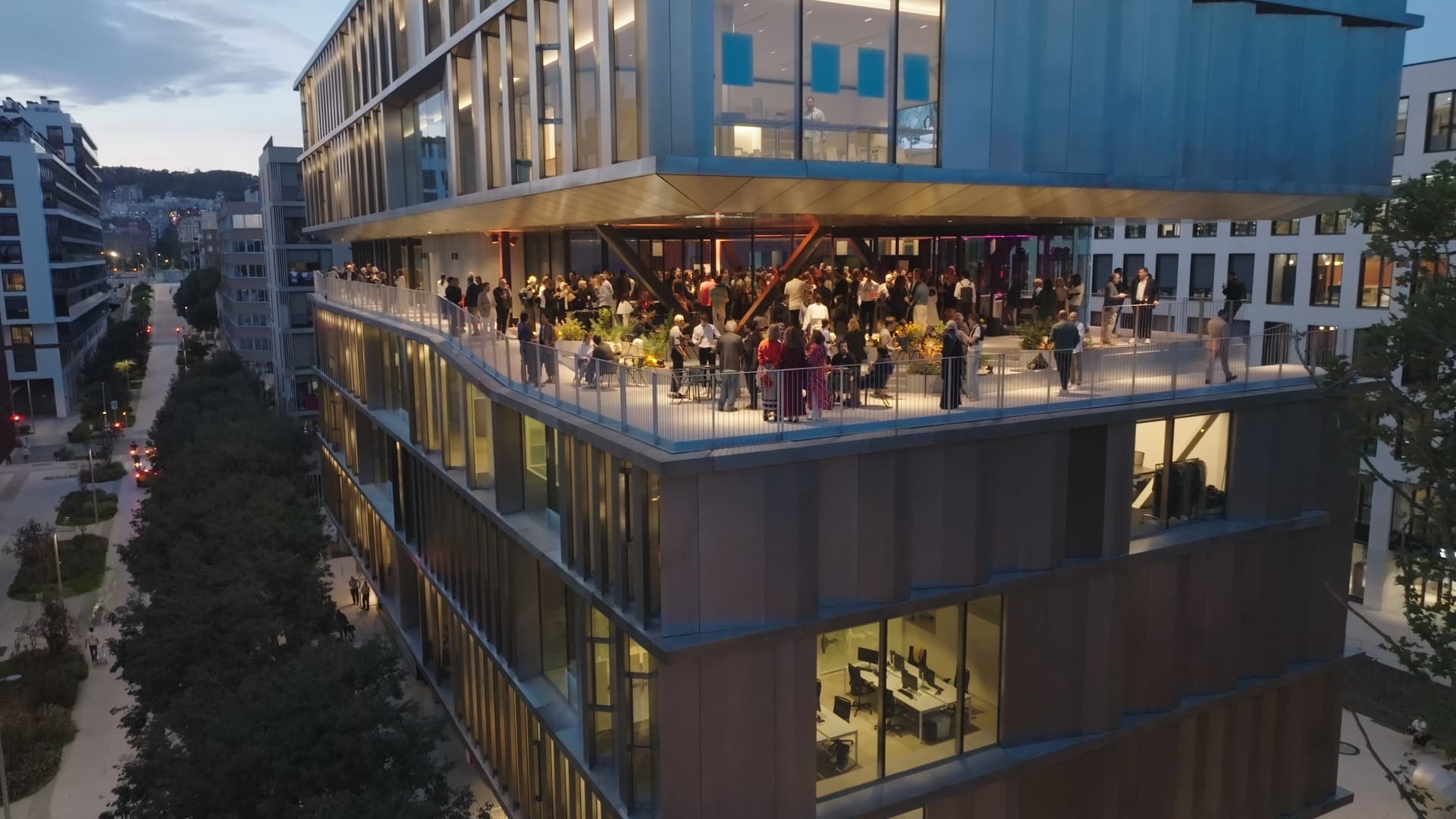 Individuals gather on a spacious terrace of a modern multi-storey office building at dusk.