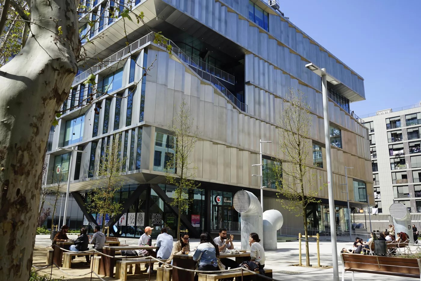 Modern glass and metal building with individuals seated at outdoor tables in a plaza on a sunny day.