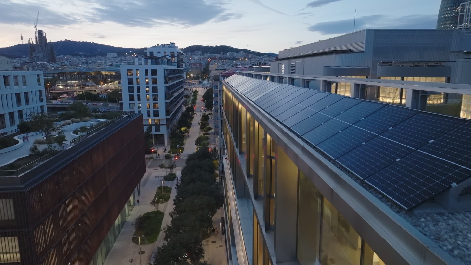 Modern city street at dusk with several buildings, including one topped with solar panels, under a softly fading sky.