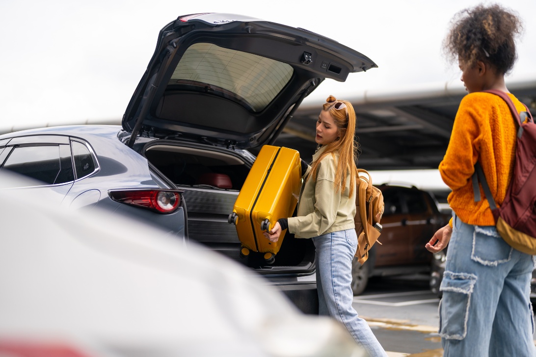 Two individuals load a yellow suitcase into the boot of a car in a car park.