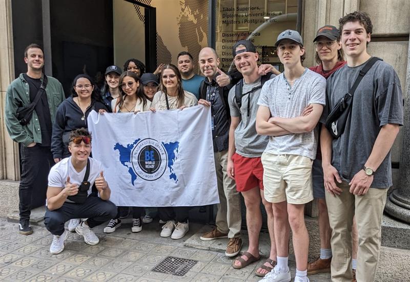 A group stands together outside a building, holding a "Be World Ready" flag and smiling at the camera.