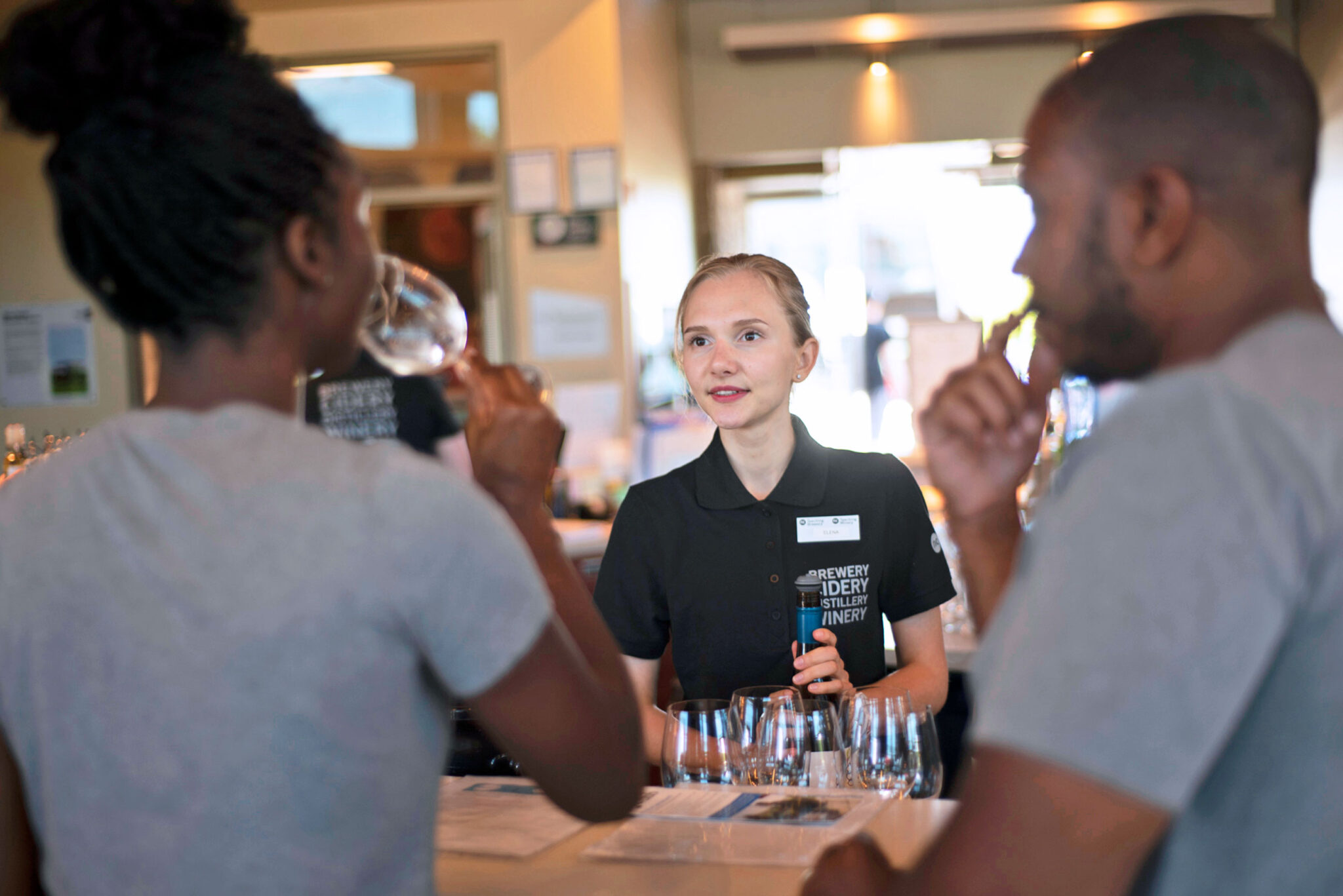 A person behind a counter talks to two customers tasting drinks at a bar or tasting room.