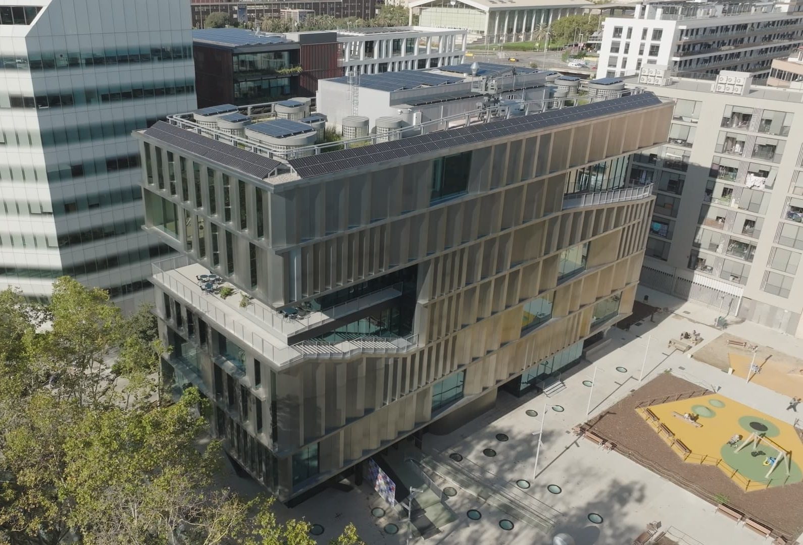 A modern, multi-storey building featuring a rooftop terrace and solar panels, surrounded by buildings and trees.