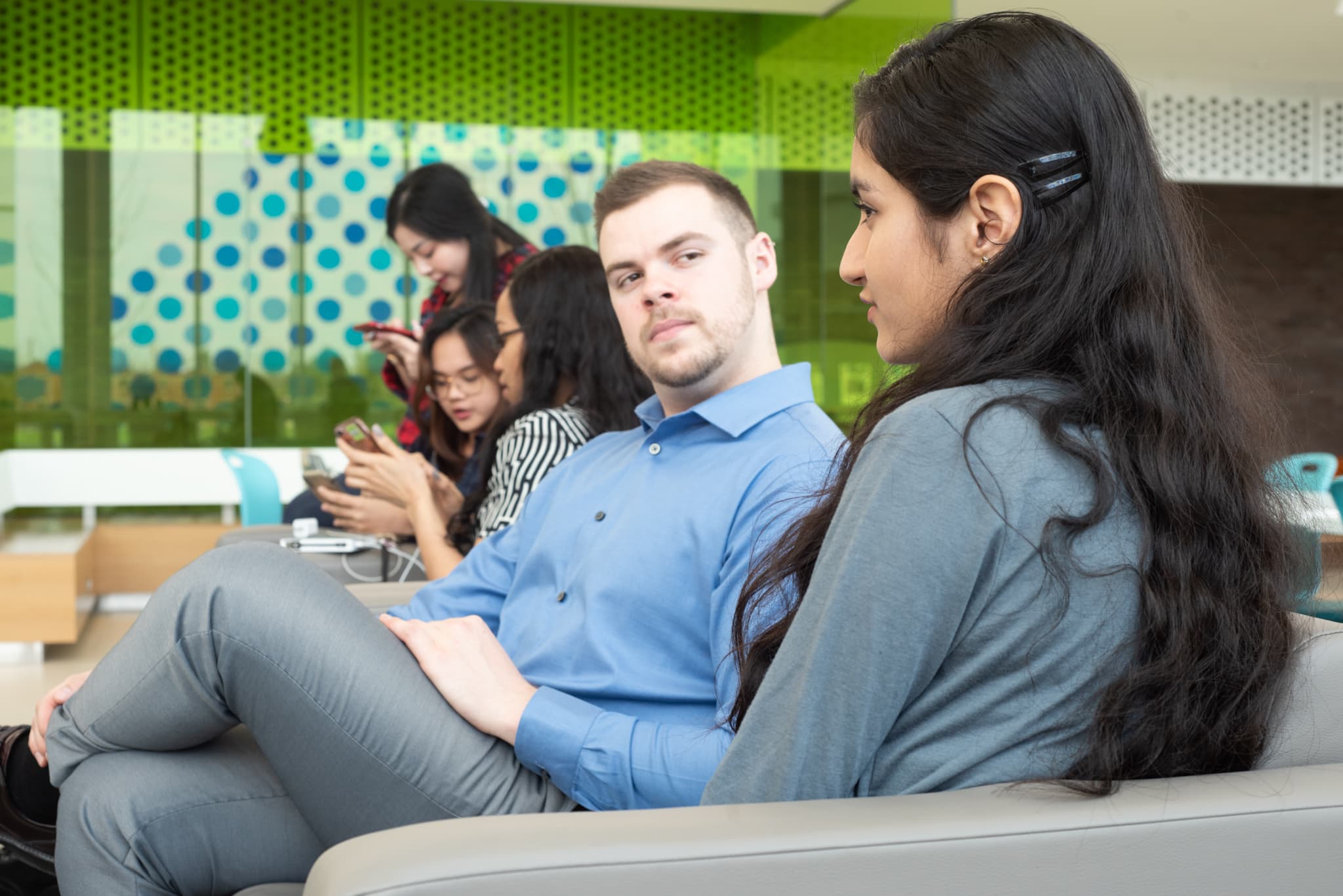 Five people sit indoors; two individuals talk in the foreground whilst three use their mobiles in the background.
