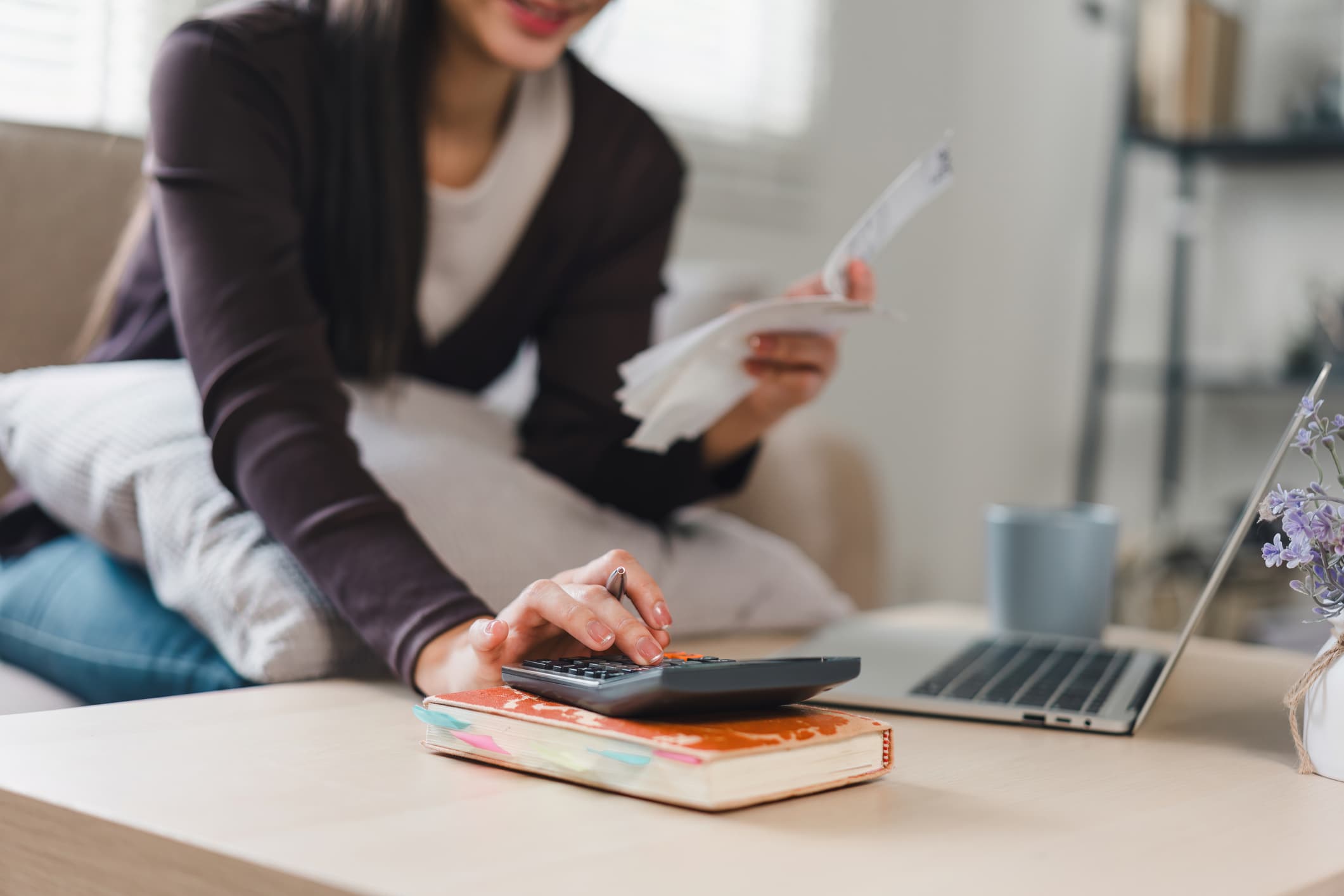 Person sitting at a table using a calculator, holding receipts, with a laptop and notebooks nearby.