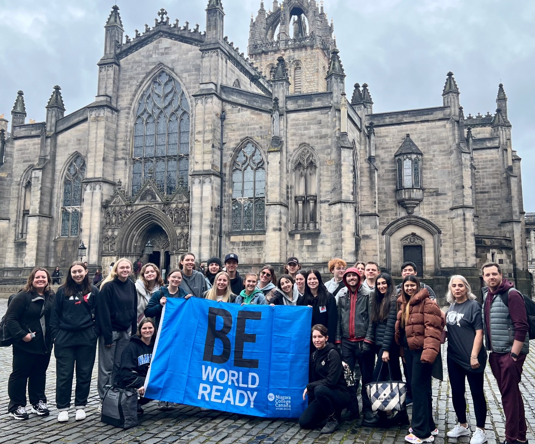 A group holding a "Be World Ready" banner stands in front of a historic stone church building.
