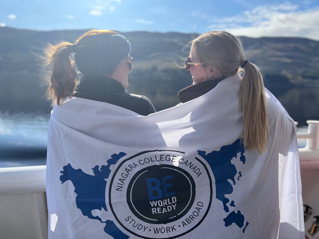 Two individuals wrapped in a Niagara College Canada “Be World Ready” flag stand outdoors by a lake, facing one another.
