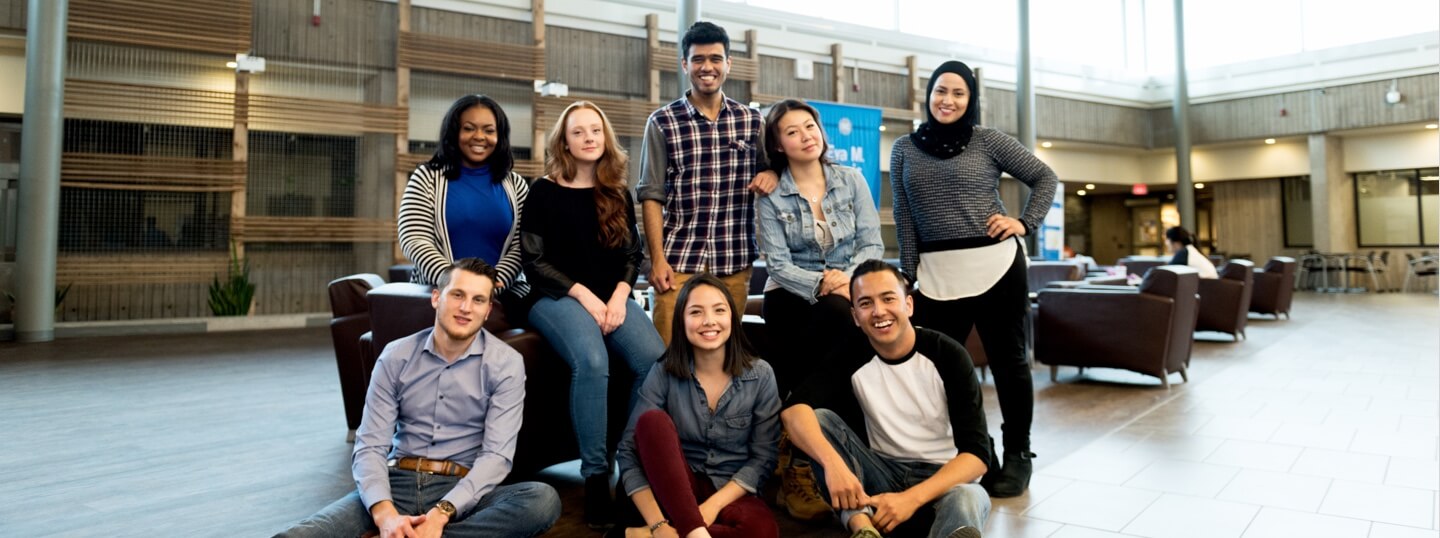 Eight young adults pose together and smile in a spacious indoor common area with chairs and tables.
