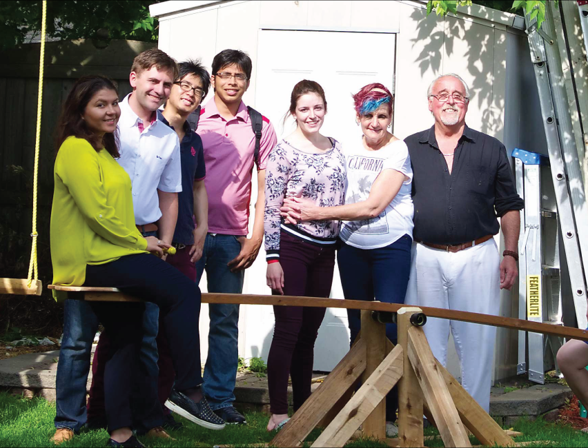 Seven individuals pose outside around a wooden see-saw in a garden, with a shed and trees visible in the background.