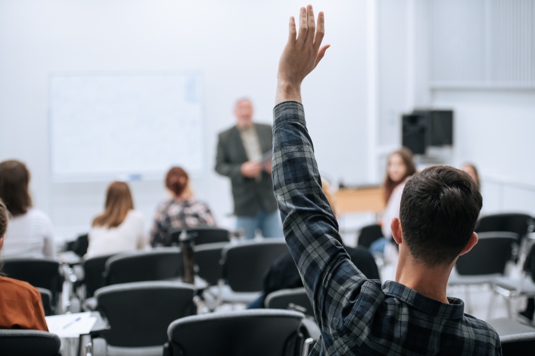 A student raises their hand to ask a question during a classroom lecture, with several people seated and a teacher at the front.