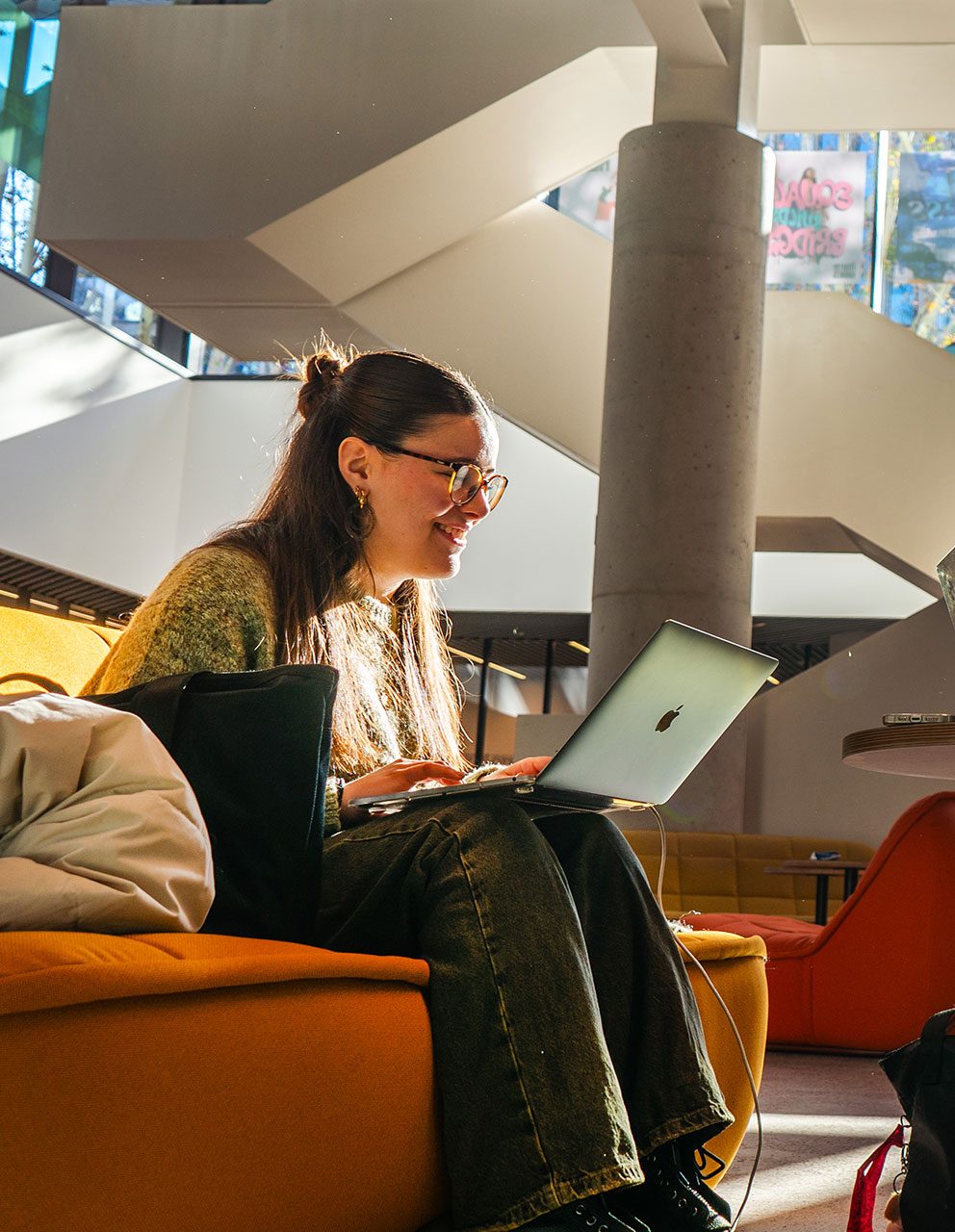 Two individuals sit opposite each other at a table in a bright lounge, both working on laptops and smiling.