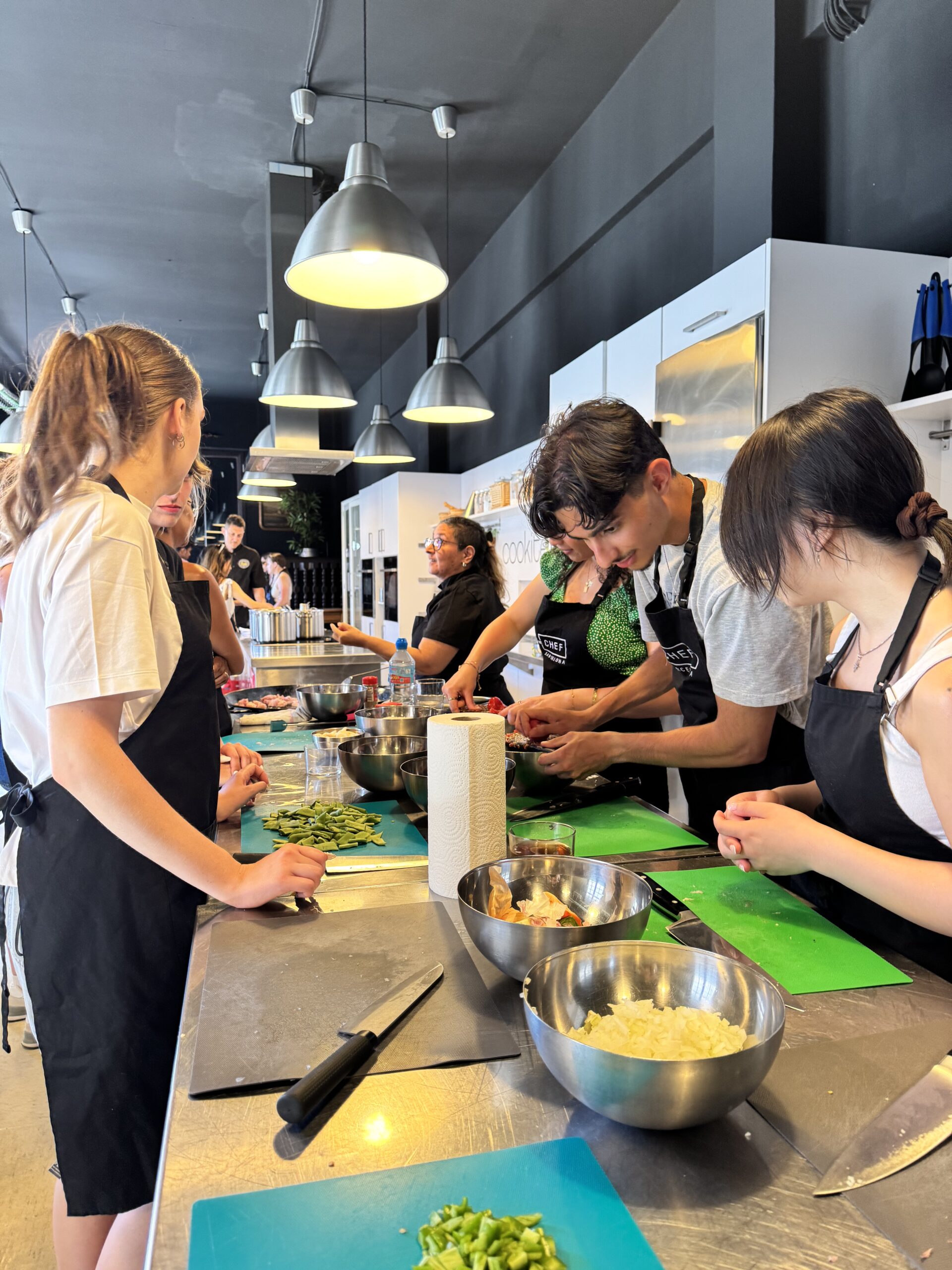 Individuals wearing aprons prepare food at a kitchen worktop during a cookery class.