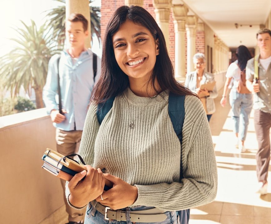 A student holding books and smiling in a school corridor, with classmates walking in the background.